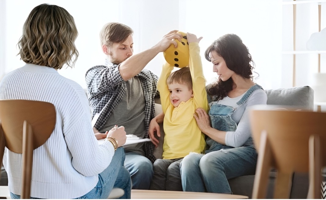 A mother and father sit on a couch together, gently assisting their young son as he holds up a large yellow plush die during a supportive family coaching session with a therapist.